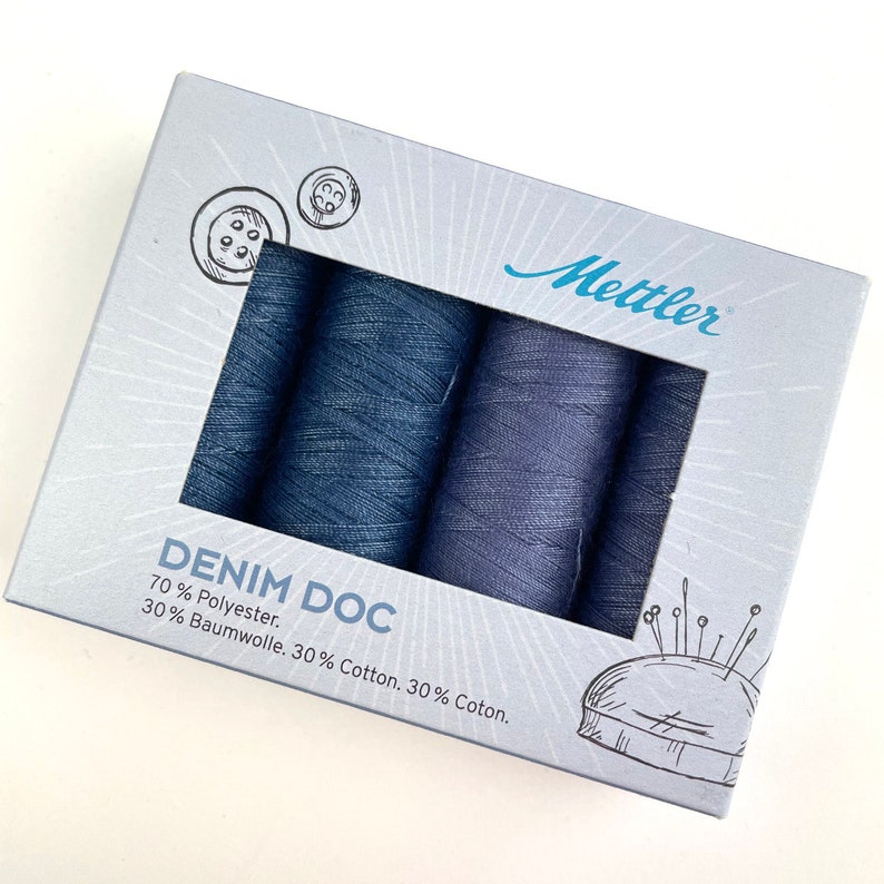 May include: A box set of Mettler Denim Doc thread spools in various shades of blue. The box is light gray with illustrations of buttons and a pincushion. The text "DENIM DOC" and thread composition are printed on the box.