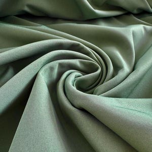 Pode incluir: Um tecido verde-sálvia sólido com acabamento fosco e uma trama fina e uniforme. O material está drapeado em dobras suaves e fluidas que criam sombras e reflexos profundos, enfatizando a textura lisa e a cor consistente do têxtil.