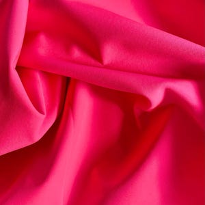 May include: Close-up of a vibrant, hot pink fabric with soft folds and subtle shadows. The material appears smooth and possibly silky, with a rich, saturated color. The fabric is bunched and draped, creating a dynamic texture.