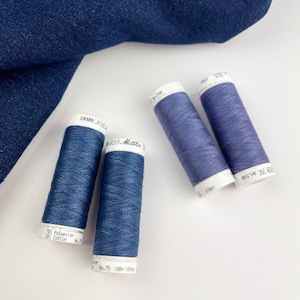 Pode incluir: Quatro carretéis de linha azul são dispostos sobre uma superfície branca, com um pedaço de tecido jeans azul ao fundo. Os carretéis de linha são rotulados com texto indicando o tipo e o comprimento da linha. A linha é azul jeans.