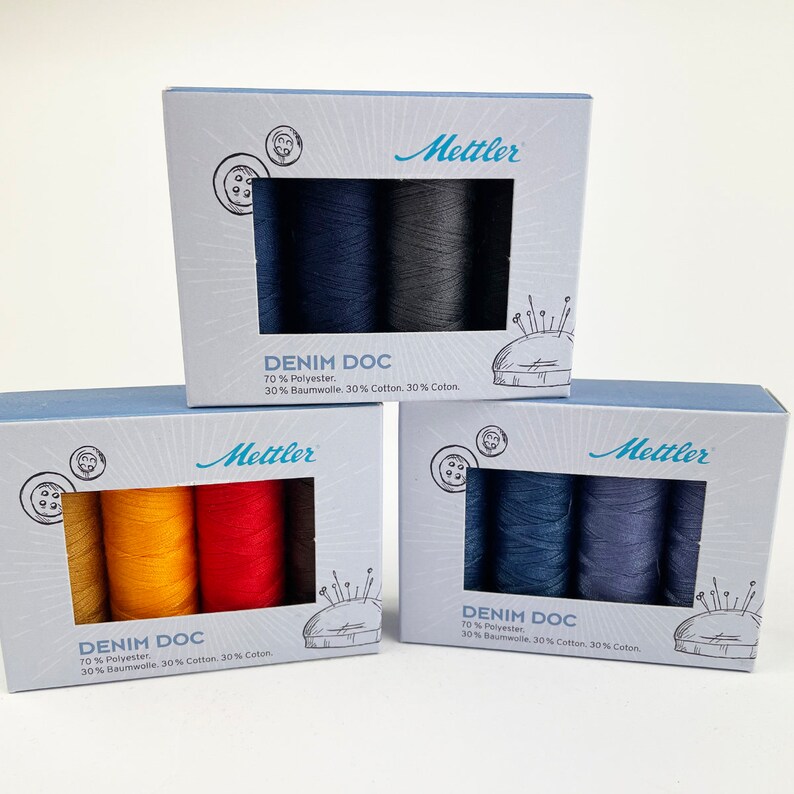 May include: Three boxes of Mettler Denim Doc thread spools. Each box has a different color scheme, including shades of blue, yellow, orange, and red. The boxes are light blue with the Mettler logo and product information printed on them.
