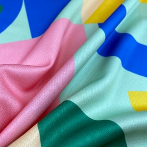 K&ouml;nnte beinhalten: Ein farbenfroher Stoff mit geometrischen Formen in Rosa, Blau, Gr&uuml;n und Gelb. Der Stoff hat eine strukturierte Oberfl&auml;che.