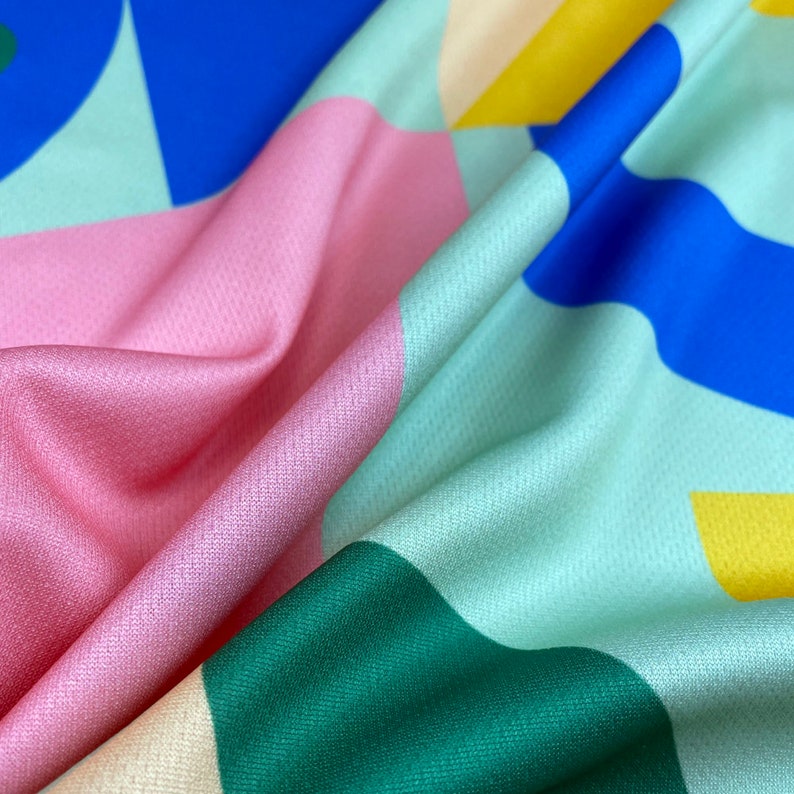 K&ouml;nnte beinhalten: Ein farbenfroher Stoff mit geometrischen Formen in Rosa, Blau, Gr&uuml;n und Gelb. Der Stoff hat eine strukturierte Oberfl&auml;che.