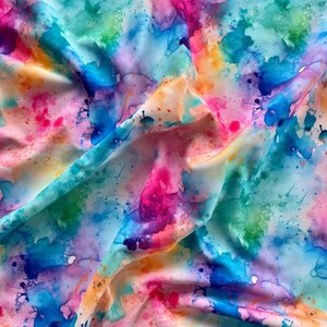 Könnte beinhalten: Ein lebendiger Stoff mit einem Aquarelldesign. Der Stoff weist eine Mischung aus Farben auf, darunter Rosa, Blau, Grün und Orange, mit einem Spritzeffekt. Der Stoff ist gefaltet, wodurch Schatten und Glanzlichter entstehen, die das Design verstärken.
