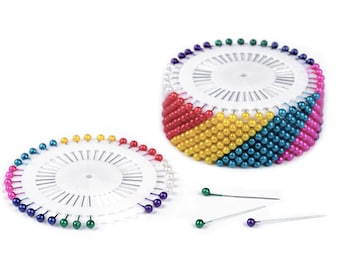 May include: A set of colorful, round-headed sewing pins in a white plastic pincushion. The pincushion has a circular base and a rotating top. The pins are arranged in a circle around the top of the pincushion. The pins are red, yellow, blue, green, and purple.