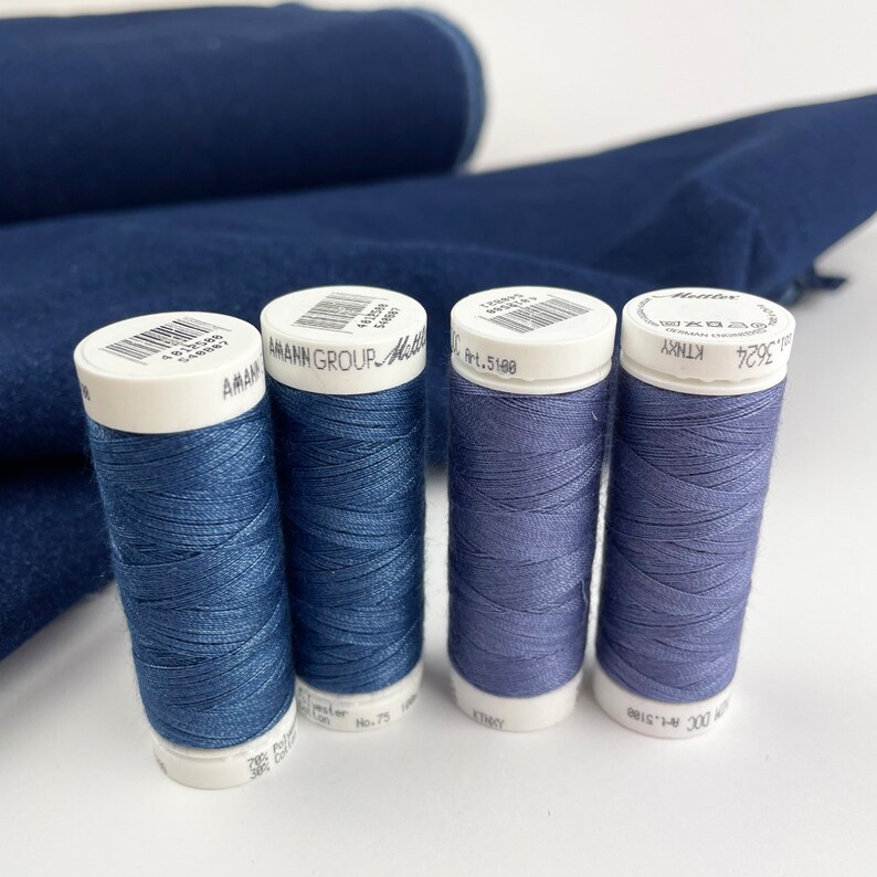 May include: Four spools of thread in shades of blue and purple, with white caps, are arranged in front of a dark blue fabric. The spools are labeled with the brand name and thread type. The fabric is folded and rolled in the background.