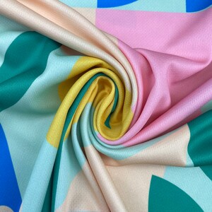 K&ouml;nnte beinhalten: Nahaufnahme eines Stoffes mit einem geometrischen Muster in Blaut&ouml;nen, Gr&uuml;n, Gelb, Rosa und Beige. Der Stoff ist gefaltet und verdreht, wodurch die verschiedenen Farben und Muster sichtbar werden.