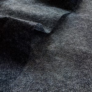 May include: Close-up of a dark gray fabric with a textured, speckled appearance. The fabric is folded, revealing layers and shadows. The material appears to be soft and possibly woven, with a subtle sheen.