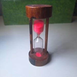 Könnte beinhalten: Eine kleine Sanduhr mit Holzrahmen und Sockel. Die Sanduhr enthält rosa Sand. Der Holzrahmen ist dunkelbraun. Die Sanduhr ist ein dekorativer Gegenstand.