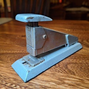 May include: A blue metal stapler with a silver top and a silver base. The stapler has a lever on the top and a metal plate on the bottom. The stapler is labeled "Swingline 303 Deluxe".