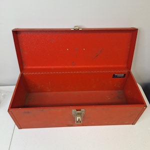 May include: Open, red metal toolbox with a hinged lid and a metal clasp. The interior is empty. The toolbox has a rectangular shape and is designed for storing tools. The brand name "Kennedy" is visible on a label inside the box.