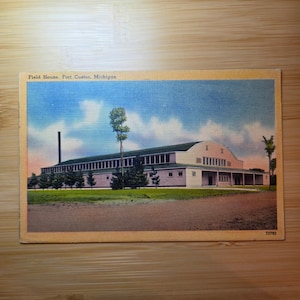 Puede incluir: Una postal antigua con una ilustración en color del Field House en Fort Custer, Michigan. El edificio tiene un techo largo y arqueado, paredes de colores claros y una chimenea alta, sobre un cielo azul con nubes y césped verde.