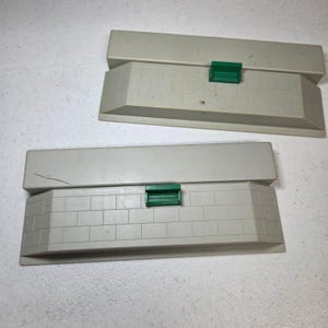 Peut inclure: Deux sections de quai de gare en plastique gris clair rectangulaires. Chaque quai présente un motif de briques moulé sur le bord avant incliné et un petit banc en plastique vert fixé au centre de la surface supérieure plate.
