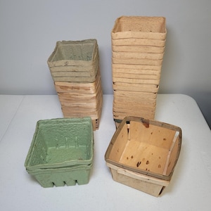 May include: A collection of square produce baskets in various materials and colors. There are stacks of light green molded fiber baskets, tan wood veneer baskets, and a single light wood basket with a dark rim, all featuring ventilation slots.