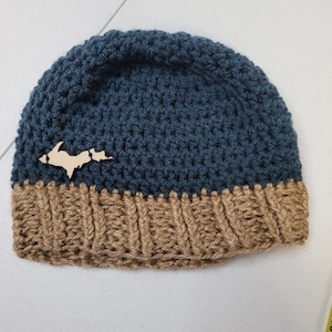 Może przedstawiać: Ręcznie robiona, niebiesko-beżowa czapka z szydełka. Górna część jest ciemnoniebieska, a rondo beżowe. Drewniane wycięcie w kształcie stanu jest przymocowane do przodu. Ta zimowa czapka jest idealna na zimną pogodę.