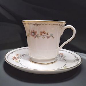 May include: A white teacup and saucer set with a floral pattern in shades of orange, yellow, and green. The cup and saucer have a gold trim around the edges.
