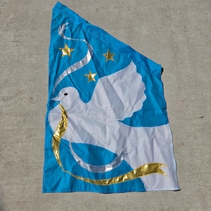 May include: A bright blue flag featuring a large white dove in the center. The dove holds a gold ribbon in its beak. Above the dove are three gold stars, and a silver ribbon curves across the upper left portion of the blue fabric background.
