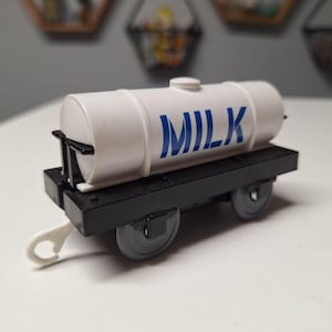 May include: A white toy milk tanker car with the word "MILK" in blue lettering. The tanker sits on a black base with gray wheels and a white hook. The toy is designed to look like a train car.