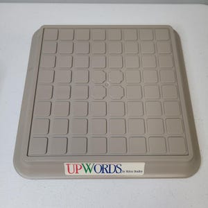 May include: Tan Upwords game board with a grid of squares. The board has a raised edge and a label that reads "UPWORDS by Milton Bradley". The game board is designed for word-building.
