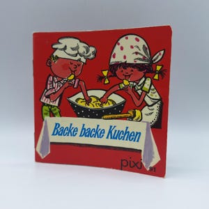 Peut inclure: Une couverture de livre pour enfants vintage avec un fond rouge. La couverture présente deux enfants de dessins animés, l'un portant une toque de chef et l'autre un foulard à pois, tous deux mangeant dans un bol. Le texte "Backe backe Kuchen" est affiché sur une bannière.