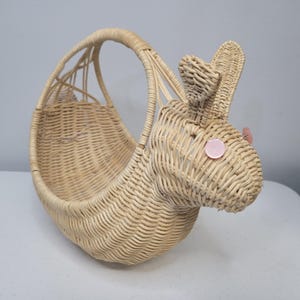 May include: A light tan woven wicker basket shaped like a rabbit. The basket features a curved handle, a rounded body, and two upright ears. Two small, round, light pink buttons are attached to the sides of the head to serve as eyes.