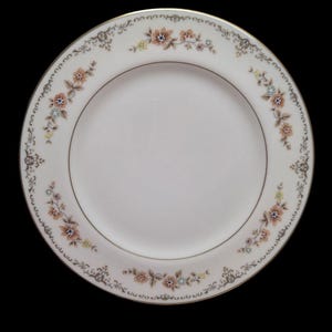 May include: White ceramic plate with a floral design. The plate features a gold rim and a border of flowers in shades of peach, blue, and yellow. The floral pattern is arranged in a repeating design around the edge of the plate.