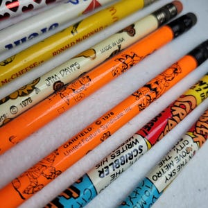 May include: A collection of wooden pencils featuring various graphic designs, including yellow pencils with Ronald McDonald and McCheese branding, orange pencils with Garfield illustrations and 1978 copyright text, and pencils with comic strip text.