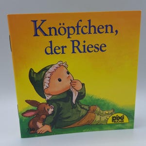 Peut inclure: Livre pour enfants intitulé "Knöpfchen, der Riese". La couverture présente une illustration de dessin animé avec un personnage en vert, un lapin brun et du texte bleu. Le fond est jaune et vert.