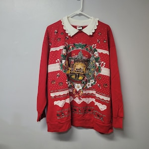 May include: A red long-sleeve sweatshirt featuring a white scalloped collar and a central graphic of a fireplace scene framed by a holiday wreath. The design includes white lace-like horizontal trim, small white dots, and festive holiday motifs.