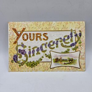 May include: A rectangular vintage postcard featuring the text "Yours Sincerely" in ornate, gold-toned lettering intertwined with purple violet flowers and green leaves. A small inset illustration depicts a landscape with a windmill and a river.