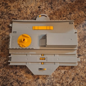 May include: A gray plastic toy train track with yellow accents. The track has a circular section, a rectangular section, and a section with a sliding switch labeled "STOP" and "GO". The track also has a yellow lever.