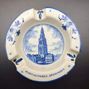 May include: Round, white ashtray with a blue and white illustration of a tall building and the text "Martinitoren Groningen". The ashtray has a blue rim and floral accents.