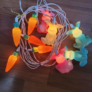 Peut inclure: Une guirlande lumineuse de Pâques avec des ampoules en forme de carottes oranges et de lapins pastel. Les lumières sont enfilées sur un fil blanc et sont allumées. Les ampoules sont dans les tons orange, jaune, rose et bleu. Les lumières sont sur une surface en bois.