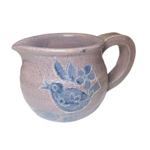 May include: A small, ceramic pitcher with a rounded body and a handle. The pitcher is a muted lavender color with a blue bird and floral design. The spout is on the left side.
