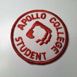 Może przedstawiać: Okrągła naszywka z haftem z napisem "APOLLO COLLEGE STUDENT" w kolorze czerwonym, otaczającym czerwony abstrakcyjny wzór na białym tle. Naszywka ma czerwoną obwódkę i jest przeznaczona na odzież lub akcesoria.