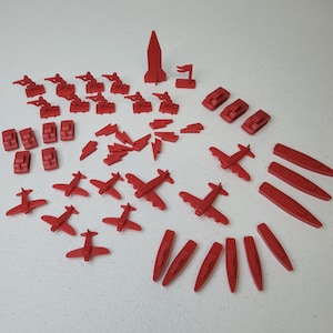May include: A collection of red plastic toy game pieces, including airplanes, tanks, boats, rockets, and soldiers. The pieces are scattered across a white surface, suggesting a board game or a child's play set.