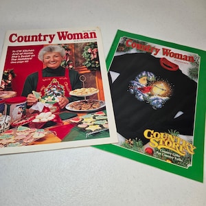 May include: Two Country Woman magazines from November/December 1998. The left magazine features a person in a red apron with holiday cookies. The right magazine is a Country Store catalog featuring a black sweatshirt with a cardinal bird graphic.