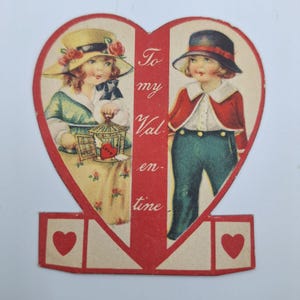 Könnte beinhalten: Vintage Valentinstagskarte in Herzform. Die Karte zeigt zwei Kinder, eines hält einen Vogelkäfig mit einem Herzen, das andere trägt einen Hut und eine Jacke. Die Karte trägt den Text "To my Valentine" in Rot.