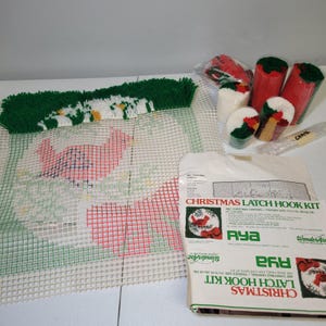 Puede incluir: Un kit de ganchillo navideño con diseño de cardenal. El kit incluye un lienzo con patrón de cuadrícula, ovillos de lana en rojo, verde y blanco, y un ganchillo. El embalaje presenta el texto "CHRISTMAS LATCH HOOK KIT".