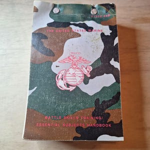 May include: A handbook with a camouflage pattern cover featuring the text "THE UNITED STATES MARINE" and "BATTLE SKILLS TRAINING/ ESSENTIAL SUBJECTS HANDBOOK". The cover also displays the emblem of the United States Marine Corps.