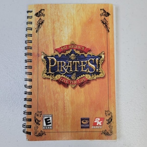 Op de afbeelding: Spiraalgebonden notitieboek met de speltitel "Sid Meier's Pirates!" op de omslag. De omslag heeft een houtnerf ontwerp met een blauw en goudkleurig embleem. De ESRB-classificatie is zichtbaar.