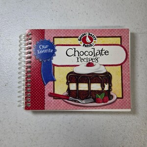 Op de afbeelding: Een spiraalgebonden kookboek getiteld Gooseberry Patch's Our Favorite Chocolate Recipes. De kaft toont een afbeelding van een chocoladetaart met witte glazuur, chocoladeganache en frambozen, naast een blauw lint-icoon.