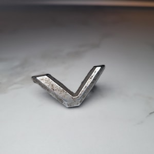 Può includere: Un emblema metallico argentato e usurato a forma di V. L'emblema presenta una superficie strutturata e segni di usura. La forma a V è l'elemento principale, su uno sfondo sfocato.
