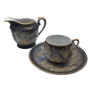 May include: A black and gold porcelain tea set featuring dragon designs. The set includes a creamer, a teacup, and a saucer. The edges of the set are trimmed with gold. The dragon designs are in relief.