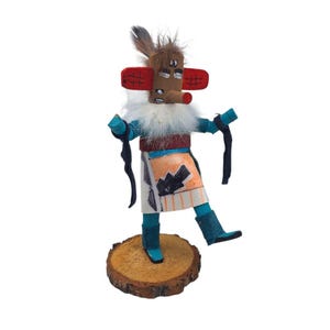 May include: A handcrafted Native American Kachina doll. The doll features a brown head with red ear-like shapes, a white beard, and a colorful outfit. It stands on a wooden base, showcasing traditional craftsmanship and cultural artistry.