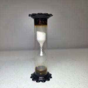 May include: An hourglass featuring a clear glass cylinder with white sand inside. The top and bottom caps are black with a scalloped, petal-like edge design. The sand is currently flowing from the top chamber into the bottom chamber of the device.