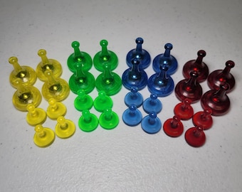 Puede incluir: Un conjunto de piezas de juego coloridas en amarillo, verde, azul y rojo. Cada pieza tiene una parte superior redondeada y una base en forma de clavija. Las piezas están dispuestas en filas, lo que sugiere un juego de mesa.