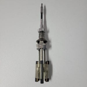 Pode incluir: Uma ferramenta de metal prateado com um corpo cilíndrico, uma mola e uma gola ajustável. Possui várias lâminas e um eixo longo e fino. A ferramenta é projetada para um propósito mecânico específico.