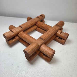 May include: A square-shaped wooden trivet constructed from interlocking cylindrical logs. The brown wood features notched grooves at regular intervals along each piece, creating a grid-like structure with a hollow centre for heat protection.