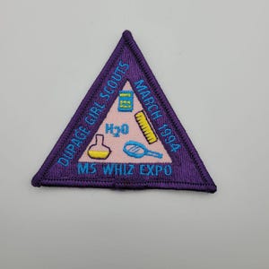 May include: A purple triangular patch with embroidered text and graphics. The text reads "DUPAGE GIRL SCOUTS", "MARCH 1994", and "MS WHIZ EXPO". Graphics include a beaker, ruler, and magnifying glass.